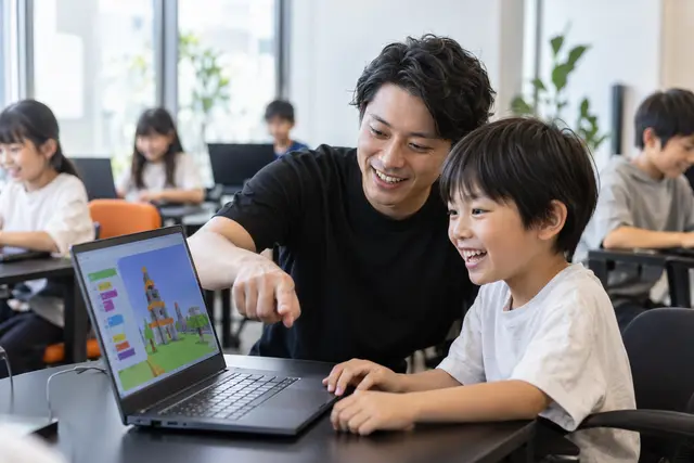 先生が子どものとなりで画面を指さし、子どもが笑っているようす（イメージ）