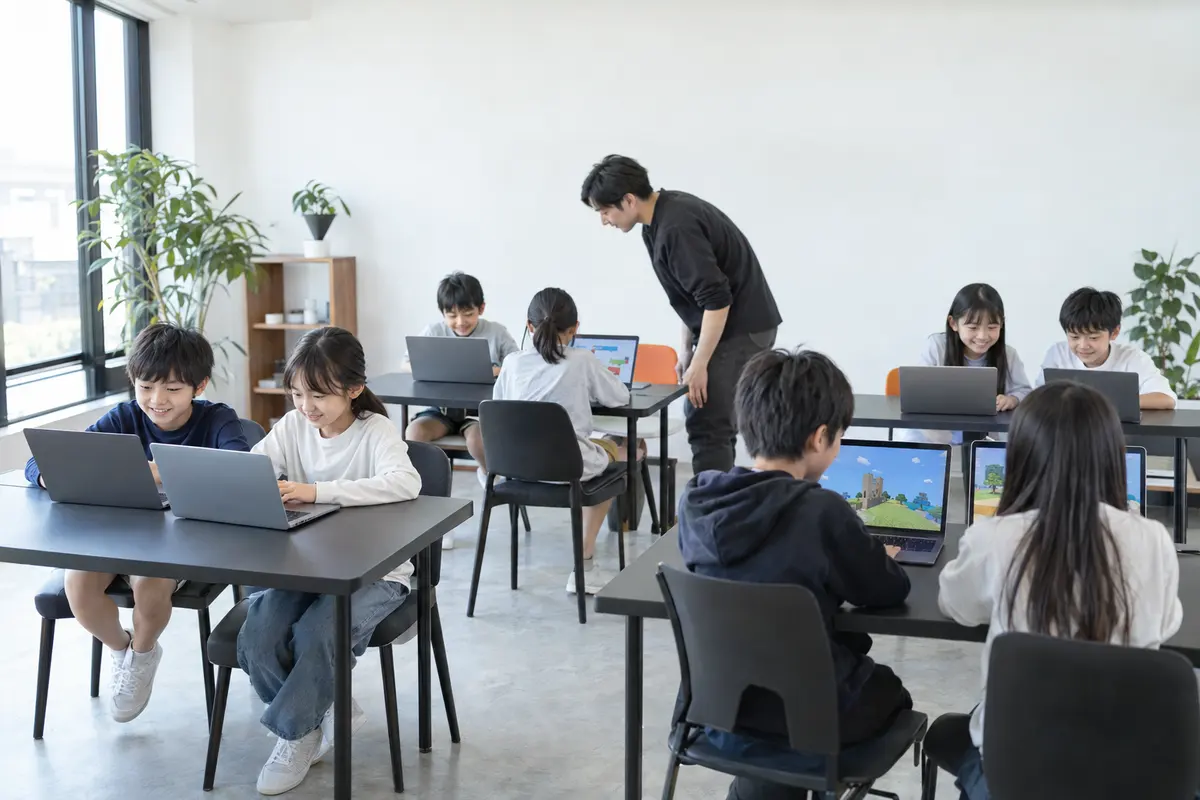 明るい教室で、子どもたちがノートパソコンに向かい、先生が席のあいだを見て回っているようす（イメージ）