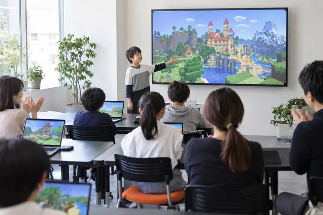 子どもが大きな画面の前で、自分のつくったゲームの世界を発表しているようす（イメージ）